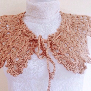 May include: A crocheted collar in a light brown color with a tie closure. The collar is made of delicate lace and has a scalloped edge.
