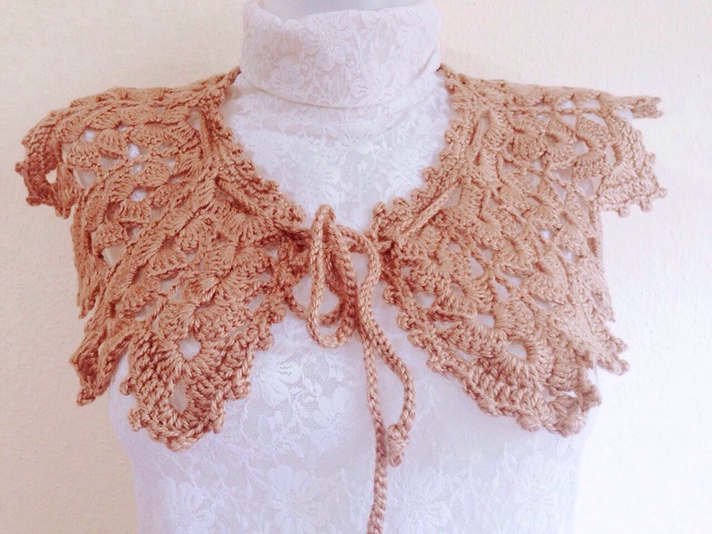 May include: A crocheted collar in a light brown color with a tie closure. The collar is made of delicate lace and has a scalloped edge.