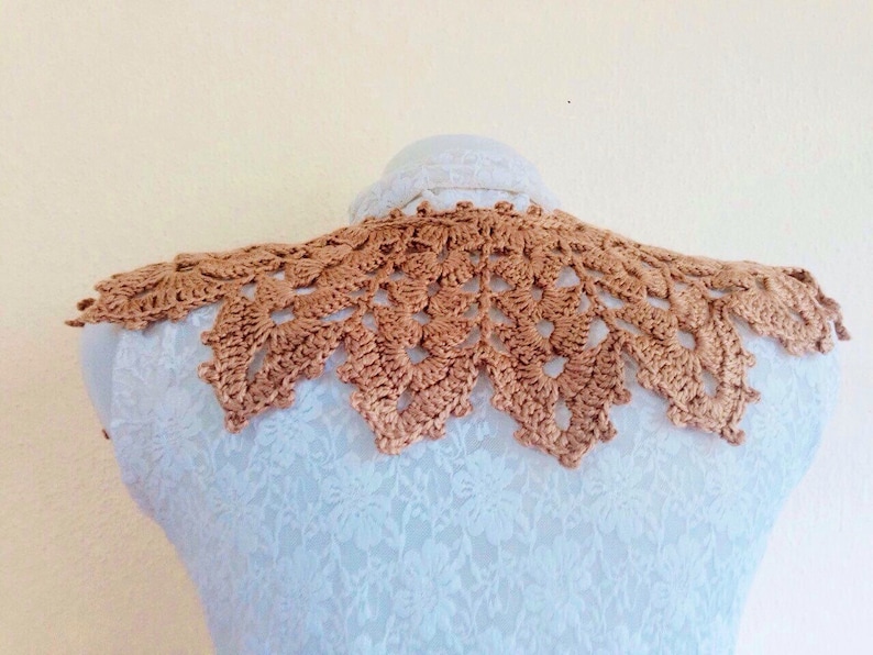 May include: A crocheted collar in a light brown color. The collar is made of delicate, intricate stitches and has a scalloped edge.