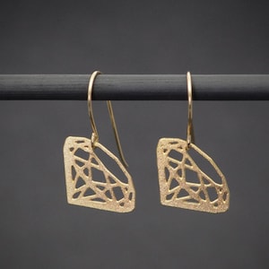 May include: A pair of gold-coloured diamond-shaped earrings with intricate geometric cutouts. The earrings hang from delicate gold hooks against a dark grey background. The earrings are displayed on a black rod.