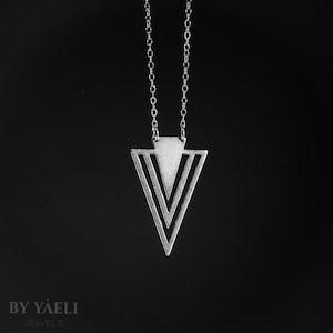 Può includere: Una collana color argento con un ciondolo triangolare dal design traforato. Il ciondolo è composto da una sezione superiore solida e due contorni a forma di V annidati, sospeso a una sottile catenina color argento su sfondo nero.