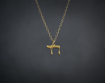 May include: A gold-toned necklace featuring a delicate chain and a small pendant shaped like the Hebrew letter Chet. The pendant hangs from a simple jump ring attached to the center of the chain, which is displayed against a dark background.