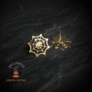 Op de afbeelding: Een paar gouden spinnenweb-oorbellen met zwarte glitteraccenten. De oorbellen hebben de vorm van een spinnenweb met een kleine spin in het midden. De oorbellen bevinden zich op een zwarte achtergrond.