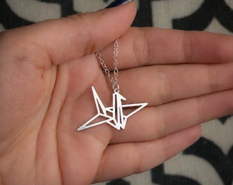 Może przedstawiać: Srebrzysty naszyjnik z geometrycznym wisiorkiem w kształcie żurawia origami. Wisiorek zawieszony jest na delikatnym srebrzystym łańcuszku. Tekst BY YAELI JEWELS jest widoczny w lewym dolnym rogu obrazu.