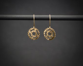 May include: A pair of gold-toned drop earrings featuring a circular gear-shaped design with a textured surface. Each earring hangs from a thin, curved wire hook. The text BY YAELI JEWELS is visible in the bottom left corner of the image.