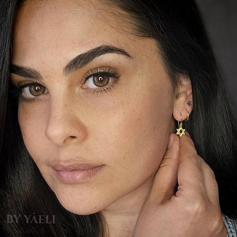 May include: A woman wearing gold earrings with a small star of David charm. The earrings are dangling from her earlobes.