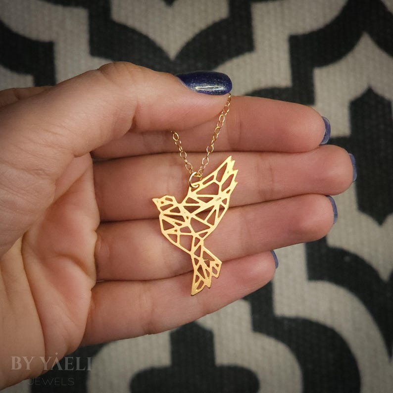 Gold-toned geometric bird pendant resting on an open hand, with a raised wing and angular cutouts visible against the skin.