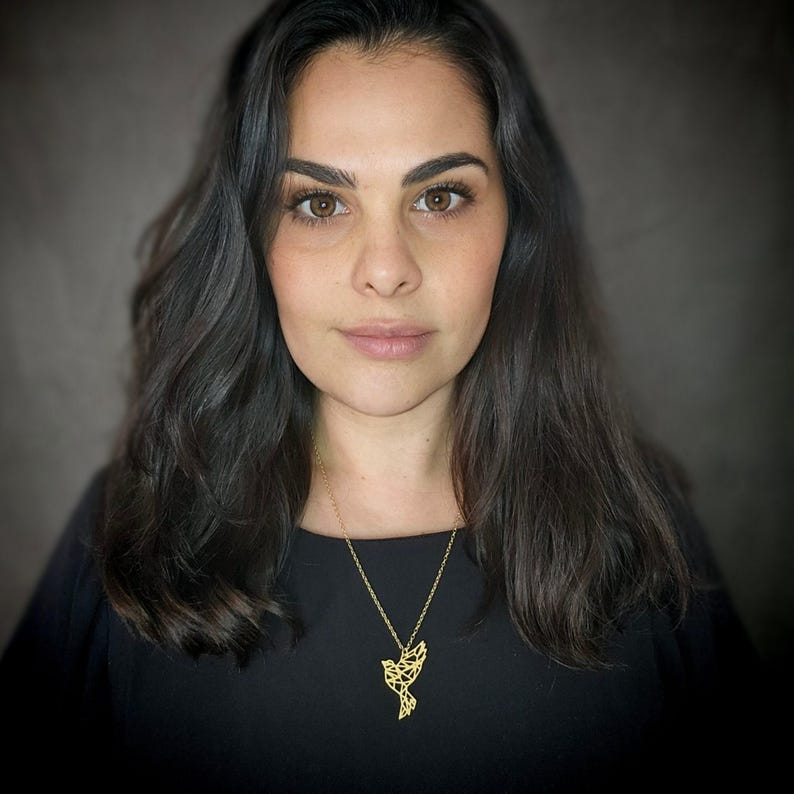 A woman in a black top wearing a gold-toned necklace with a geometric bird pendant, shown in a front portrait.