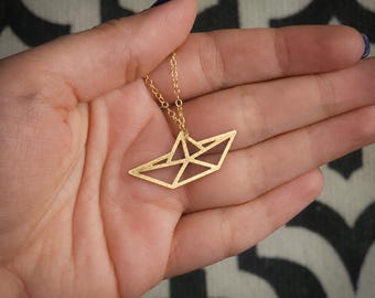 May include: A gold-toned necklace featuring a pendant shaped like a geometric paper boat. The pendant has a textured, matte finish and is suspended from a delicate gold-toned chain. The text BY YAELI JEWELS is visible in the bottom corner.