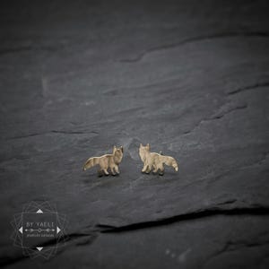 May include: A pair of gold-toned stud earrings shaped like foxes with a hammered, textured finish. The earrings feature a stylized silhouette of a standing fox with a bushy tail. A logo with the text BY YAELI JEWELRY DESIGN is visible.