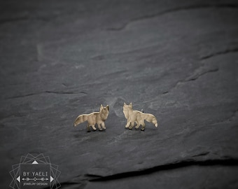 May include: A pair of gold-toned stud earrings shaped like foxes with a hammered, textured finish. The earrings feature a stylized silhouette of a standing fox with a bushy tail. A logo with the text BY YAELI JEWELRY DESIGN is visible.