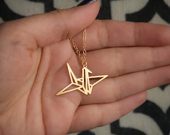 Könnte beinhalten: Eine goldfarbene Halskette mit einem geometrischen Origami-Kranich-Anhänger an einer feinen Kette. Der Anhänger ist als stilisierter Vogel mit scharfen, kantigen Linien geformt. Der Text BY YAELI JEWELS ist in der unteren Ecke sichtbar.