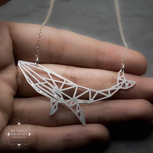 Silver-tone openwork whale pendant held across the fingers, showing its angular body, fins and upturned tail.