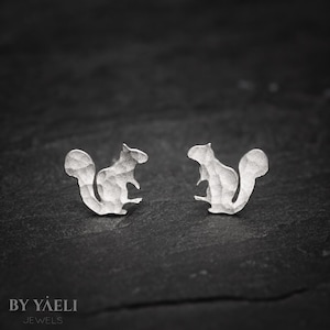 May include: A pair of silver stud earrings shaped like squirrels. Each earring features a textured, hammered metal surface with a distinct silhouette of a squirrel in a seated position, facing inward toward the other earring in the pair.