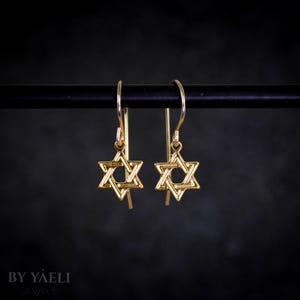 May include: A pair of gold-toned dangle earrings featuring a Star of David design. The earrings are on a black stand.