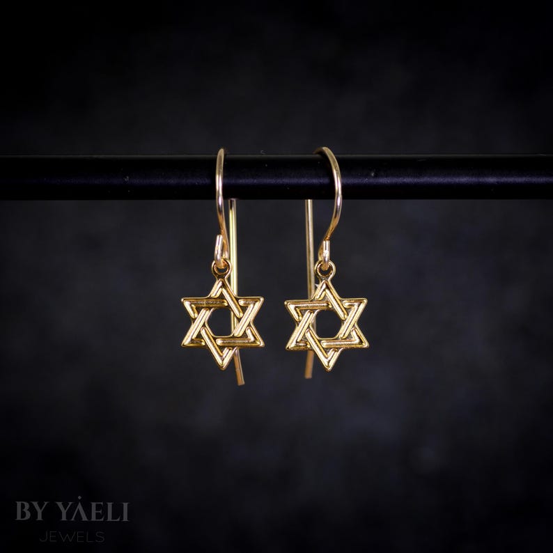 May include: A pair of gold-toned dangle earrings featuring a Star of David design. The earrings are on a black stand.