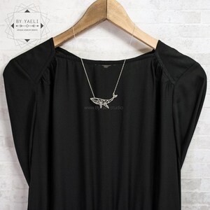 Silver-tone geometric whale necklace displayed on a black blouse hanging from a wooden hanger against white brick.