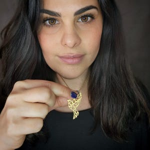 A woman holds a gold-toned openwork bird pendant near her neckline, showing its pointed wing and tail.