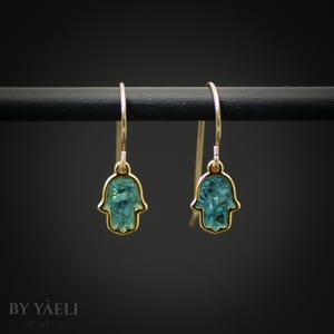 May include: A pair of gold-toned drop earrings featuring a Hamsa hand charm with a textured, teal-colored inlay. The earrings hang from thin gold-toned hooks. The text BY YAELI JEWELS is visible in the bottom left corner of the image.