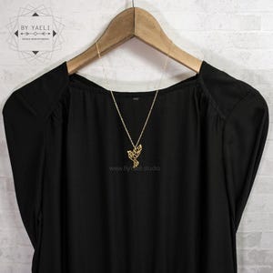 Gold-toned bird necklace draped over a black blouse on a wooden hanger against a white brick wall.