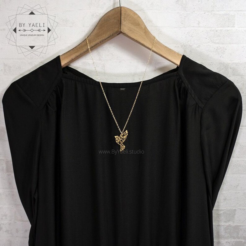 Gold-toned bird necklace draped over a black blouse on a wooden hanger against a white brick wall.