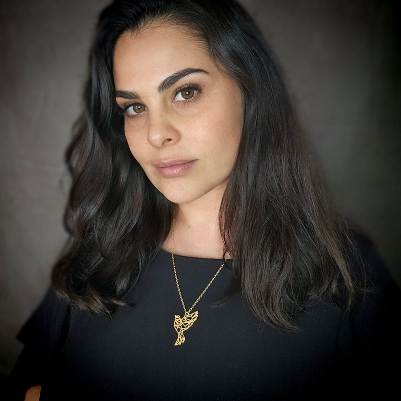 A woman wearing a gold-toned geometric bird necklace over a black top, photographed with her head slightly turned.