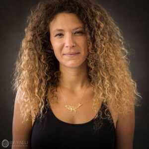 Woman with curly hair wearing a small gold-tone geometric whale necklace above a black tank top.