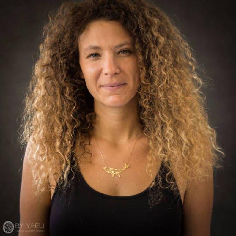 Woman with curly hair wearing a small gold-tone geometric whale necklace above a black tank top.