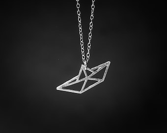May include: A silver-toned necklace featuring a pendant shaped like a geometric paper boat. The pendant has a textured, matte finish and hangs from a delicate silver-toned chain. The text BY YAELI JEWELS is visible in the bottom left corner.