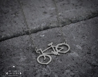 Könnte beinhalten: Eine silberfarbene Halskette mit einem fahrradförmigen Anhänger an einer feinen Kette. Der Anhänger ist ein minimalistischer Umriss eines Fahrradrahmens mit zwei runden Rädern. Der Text BY YAELI ist in der unteren linken Ecke sichtbar.