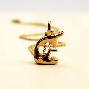 May include: A gold-colored microscope pendant necklace. The pendant is a detailed miniature of a microscope, with a chain and clasp. The necklace is displayed on a light-colored surface.