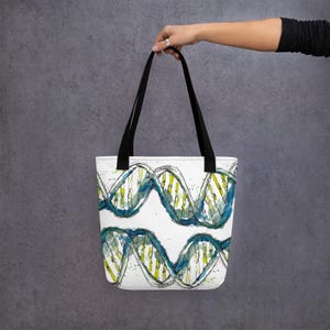 Puede incluir: Una bolsa de tela blanca con asas negras que presenta una ilustración de acuarela de una doble hélice de ADN en tonos azules, verdes y amarillos.