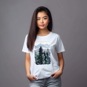 Könnte beinhalten: Weißes T-Shirt mit einem Aquarelldruck einer Berglandschaft mit grünen Bäumen und blauem Himmel.