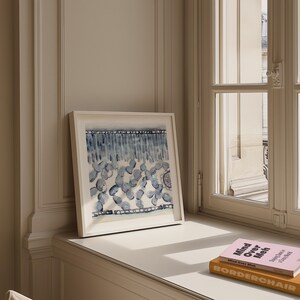 Op de afbeelding: Een ingelijste aquarel met een witte rand, met blauwe en witte abstracte vormen. Het kunstwerk wordt op een vensterbank naast twee boeken tentoongesteld. Het ontwerp van het schilderij omvat verticale en afgeronde vormen.