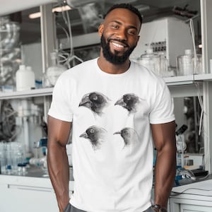 Puede incluir: Camiseta blanca con cuatro ilustraciones en escala de grises de cabezas de pájaros. La obra de arte muestra diferentes ángulos del pico y el ojo del pájaro. La camiseta es de cuello redondo clásico.