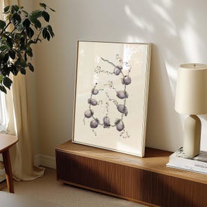 May include: A framed art print featuring a watercolor illustration of a necklace. The necklace design has a series of purple, rounded stones connected by a delicate chain. The artwork is set against a cream-colored background and framed in a gold-toned frame.