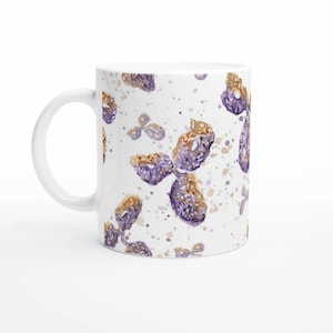 Könnte beinhalten: Weiße Keramik-Kaffeetasse mit einem Aquarelldesign aus lila und goldenen Blumenformen und gesprenkelten Akzenten. Die Tasse hat einen gebogenen Griff und eine glänzende Oberfläche, geeignet für heiße oder kalte Getränke.