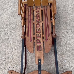 May include: A vintage wooden sled featuring a high backrest with a cartoon character graphic, a seat with the text SPEEDAWAY in red, and a front steering bar labeled GLADDING. The sled has black metal runners and side supports with wood slats.