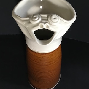 May include: A white ceramic pitcher with a whimsical face, featuring large eyes and an open mouth. The pitcher has a brown glazed base.