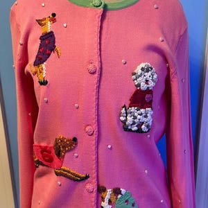 May include: Pink cardigan sweater with green trim and button closure. The sweater features embroidered dog designs wearing sweaters and coats, with heart-shaped embellishments and small pearl accents.