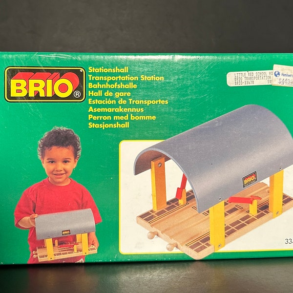 May include: A green box with a picture of a child holding a wooden train station. The box has the text "BRIO" and "Stationshall" on it. The train station is grey and yellow and has a curved roof.