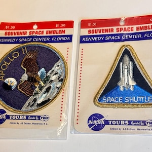 May include: Two souvenir space emblems from Kennedy Space Center, Florida. One is a circular patch with an eagle and the text "APOLLO II". The other is a triangular patch with a space shuttle and the text "SPACE SHUTTLE". Both are on a clear plastic backing.