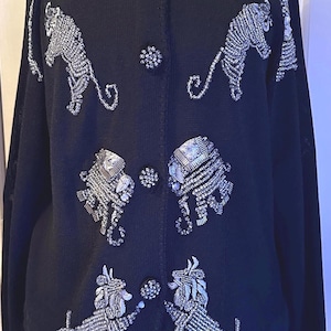 May include: Black cardigan sweater with silver beaded poodle designs. The sweater has a Peter Pan collar and decorative buttons. The poodle designs are on the front and sleeves of the sweater. The sweater is displayed on a mannequin.