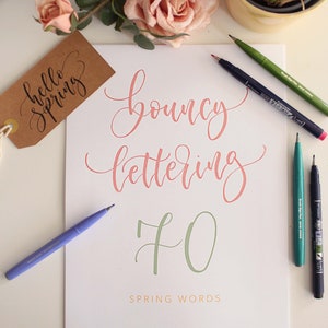 Puede incluir: Una hoja blanca con caligrafía coral y verde que dice "bouncy lettering" y "70 spring words". Una etiqueta marrón con "hello spring" está a la izquierda. Bolígrafos y rosas rosas están dispuestos alrededor.