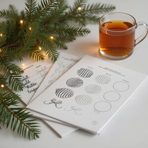May include: A clear glass mug filled with tea sits next to a stack of white paper with calligraphy practice exercises. A sprig of evergreen with small lights is in the background.