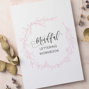 Op de afbeelding: Een witte werkmap met de tekst "Mindful Lettering Workbook" in sierlijke letters, omcirkeld door een delicate roze bloemenkrans. De cover is strak en minimalistisch, wat duidt op creatieve oefening en mindfulness.