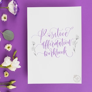 Pode incluir: Um caderno branco com as palavras "Positive Affirmation Workbook" em escrita roxa. O caderno é rodeado por flores brancas e roxas sobre um fundo roxo. O caderno tem um design floral.