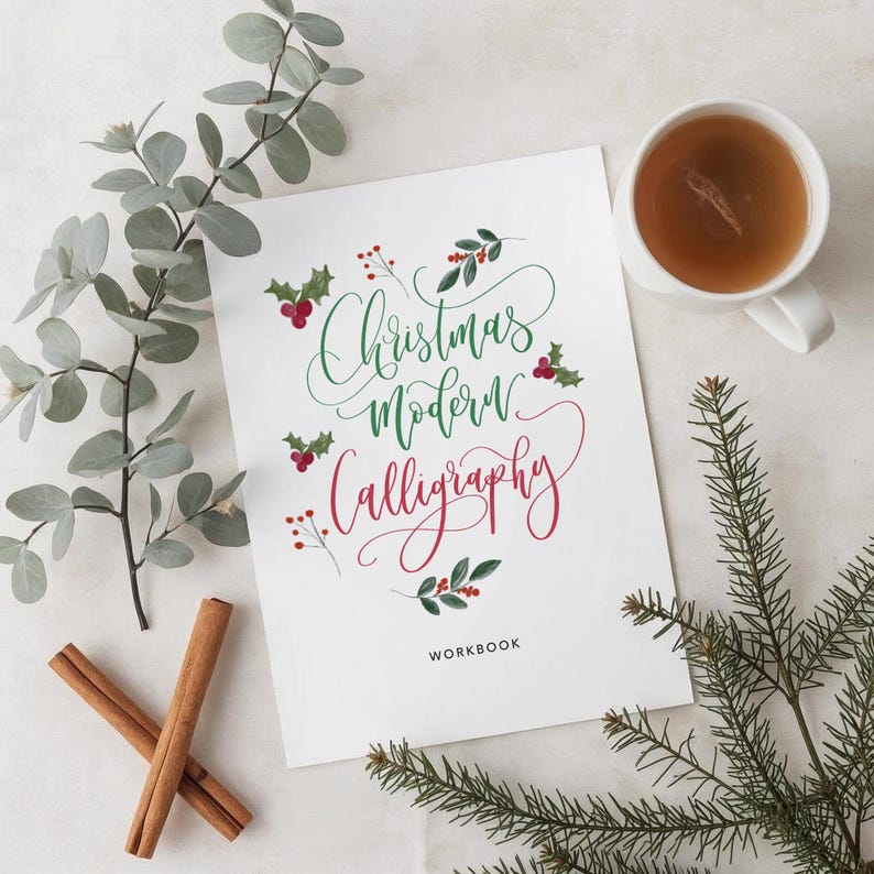 May include: A white workbook with the words "Christmas Modern Calligraphy" in green and red script, decorated with holly and berries. Cinnamon sticks, a cup of tea, and greenery surround the workbook.