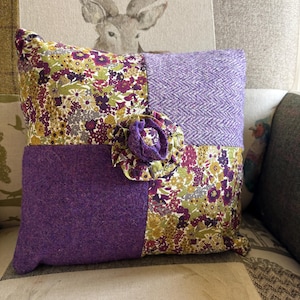 May include: A decorative square pillow with a patchwork design. The pillow features sections of floral fabric, herringbone weave, and solid purple wool. A large, ruffled purple fabric flower adorns the front. The pillow is placed on a patterned sofa.