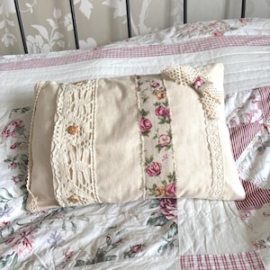 May include: Cream rectangular cushion featuring floral and lace embellishments. A central strip displays pink roses and green leaves. The cushion is trimmed with cream lace and decorated with small buttons.